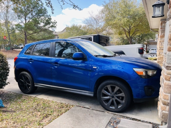 Blue Mitsubishi Outlander SUV sitting in a residential driveway in Houston, TX, showing an as-is vehicle that can be picked up with free towing.