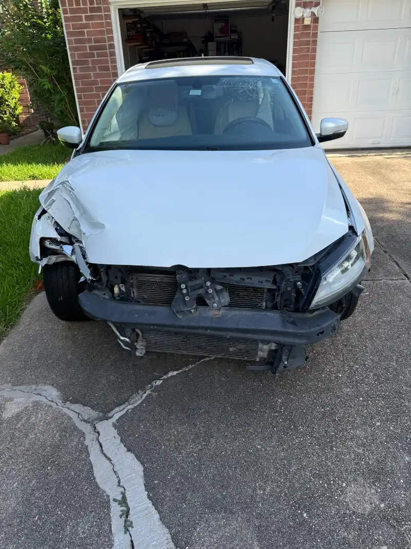 White 2015 Volkswagen Jetta with front-end collision damage and a cracked windshield parked in a Jersey Village driveway.